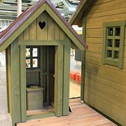 Bilden föreställer ett litet grönt utedass. Ovanför ingången är det ett utsågat hjärta i väggen. I bakgrunden syns en lokal med flera hus under uppbyggnad. 