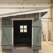 Fotografiet föreställer en verkstad. Verkstaden är grön och har ett snedtak. Husets knutar är vita. Dörrarna till verkstaden är öppna och igenom dörrarna ser man ett fönster med spröjs. 