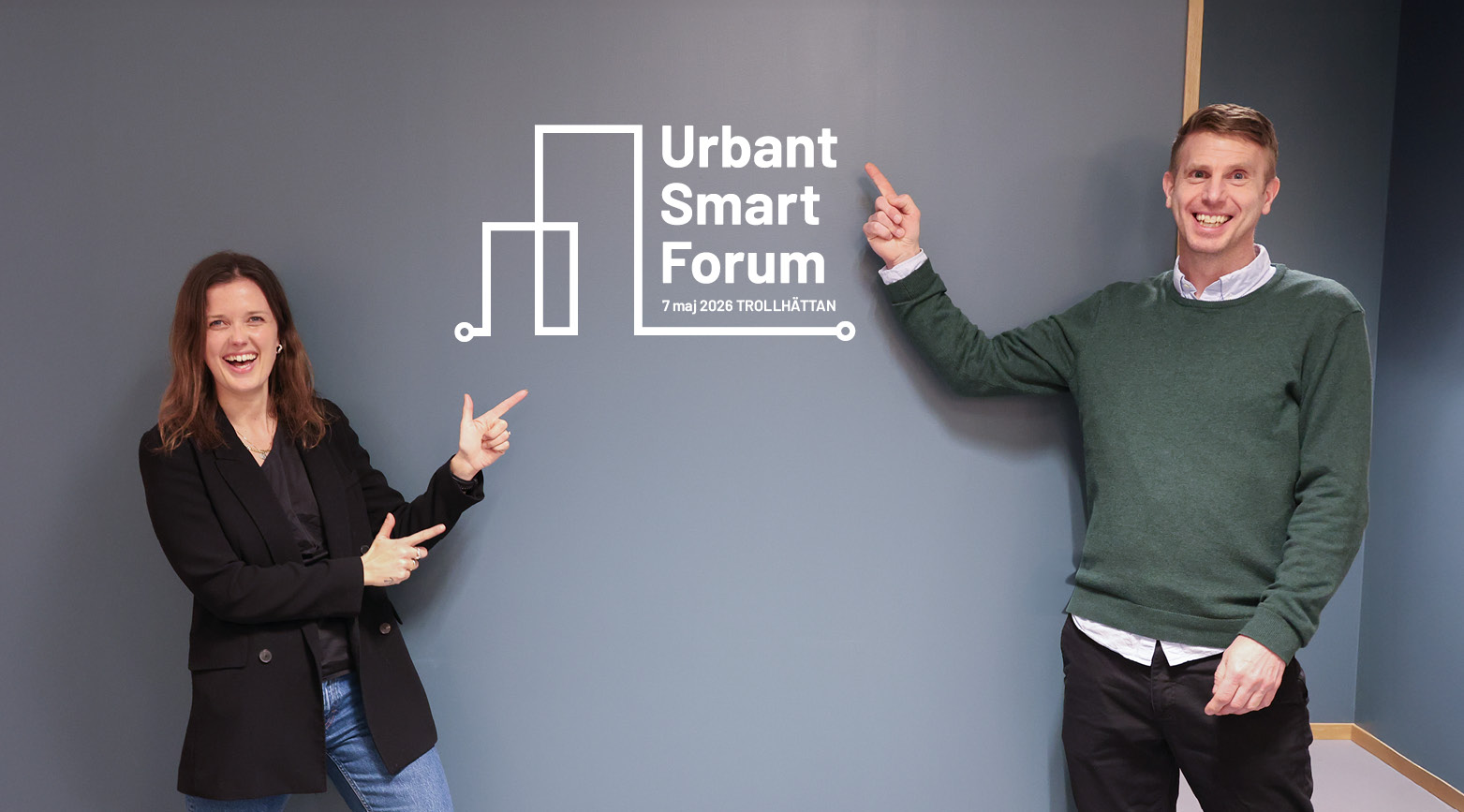 Hanna Marke och Fredrik Sandberg pekar på väggen där det står "Urbant Smart Forum". 