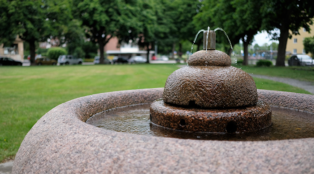 Fotografiet är en närbild på en fontän i sten. I bakgrunden syns en gräsmatta och flera stora träd som står på rad.