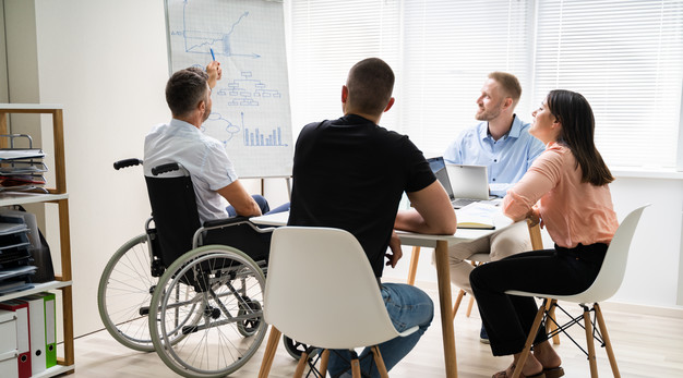 Person i rullstol sitter på ett möte och håller i en presentation för tre andra personer som sitter vid ett bord.