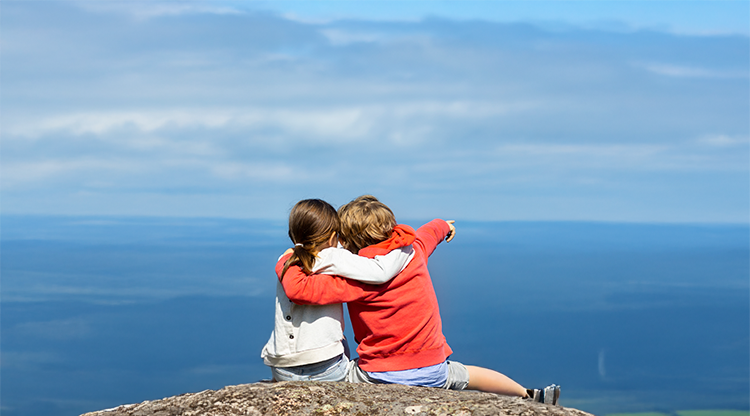 Två barn sitter på ett berg och tittar ut över havet. Man ser deras ryggar, havet och en blå himmel. 