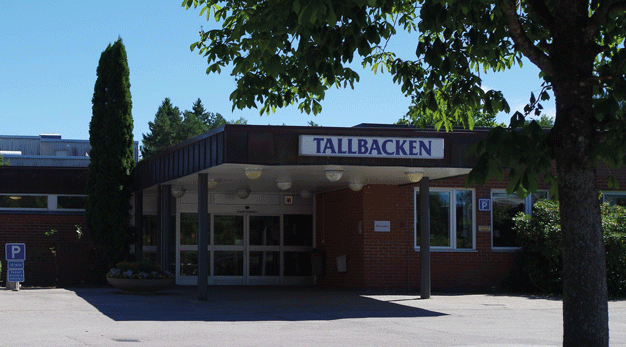 En stort träd står på en liten gräsyta i mitten av en vändplan framför en byggnad. På byggnaden sitter en stor skylt där det står Tallbacken. Under skylten är en ingång gjord av glasdörrar.