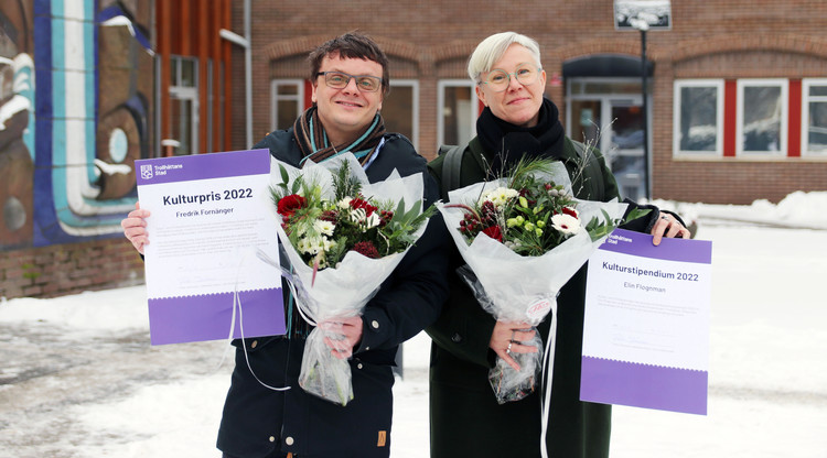 Två personer står utomhus med blomma och diplom.