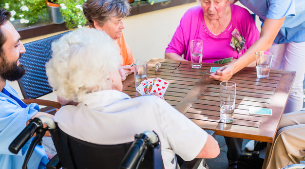 Ett gäng äldre personer sitter och spelar kort utomhus runt ett bord.