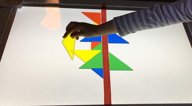 Bilden föreställer trianglar som ligger på ett bord som lyser. Trianglarna är i olika färger så som gula, blåa, röda och gröna. Från höger i bild syns en barnhand som placerar en gul triangel på bordet. 