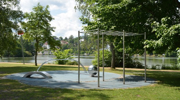 Fotografiet föreställer Spiköns utegym. I bild syns en ställning för armgång, en stor boll och en ställning för dips. Alla redskapen är i silver och i bakgrunden syns älven. Runt utegymmet syns grönska.  