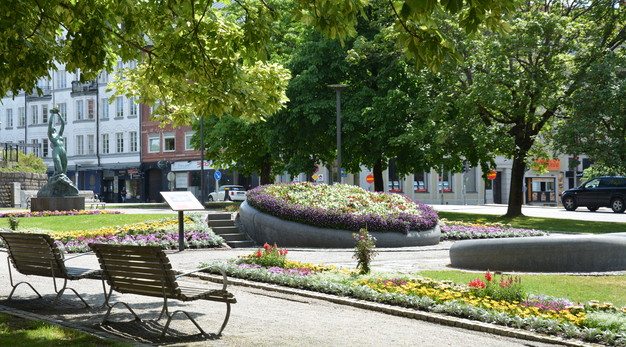 Fotografiet föreställer en park. I mitten av parken är det en stor rund blomsterrabatt med flera olika typer a blommor. Längst kanten på rabatten är det lila blommor. I bakgrunden syns konstverket den heliga lågan, hus och flera gröna stora trädkronor. I förgrunden syns ryggen av två parkbänkar och en grusgång. Fotografiet är taget på sommaren och det riktigt lyser om alla blommor.