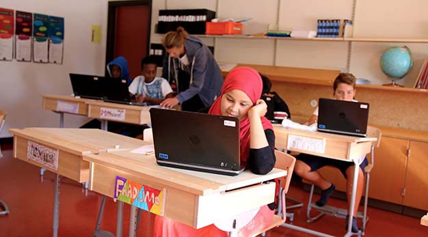 Bilden föreställer en flicka som sitter vid sin skolbänk och tittar ner i sin dator. Flickan ler. I bakgrunden sitter flera barn och studerar. En lärare står vid en av eleverna och pekar på ett papper. 