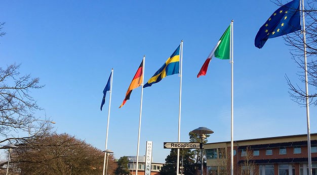 Bilden föreställer fem flaggstänger med två Eu-flaggor, Tysklands flagga, Sveriges flagga och Italiens flagga. Bilden är tagen utanför Trollhättans stadshus. 
