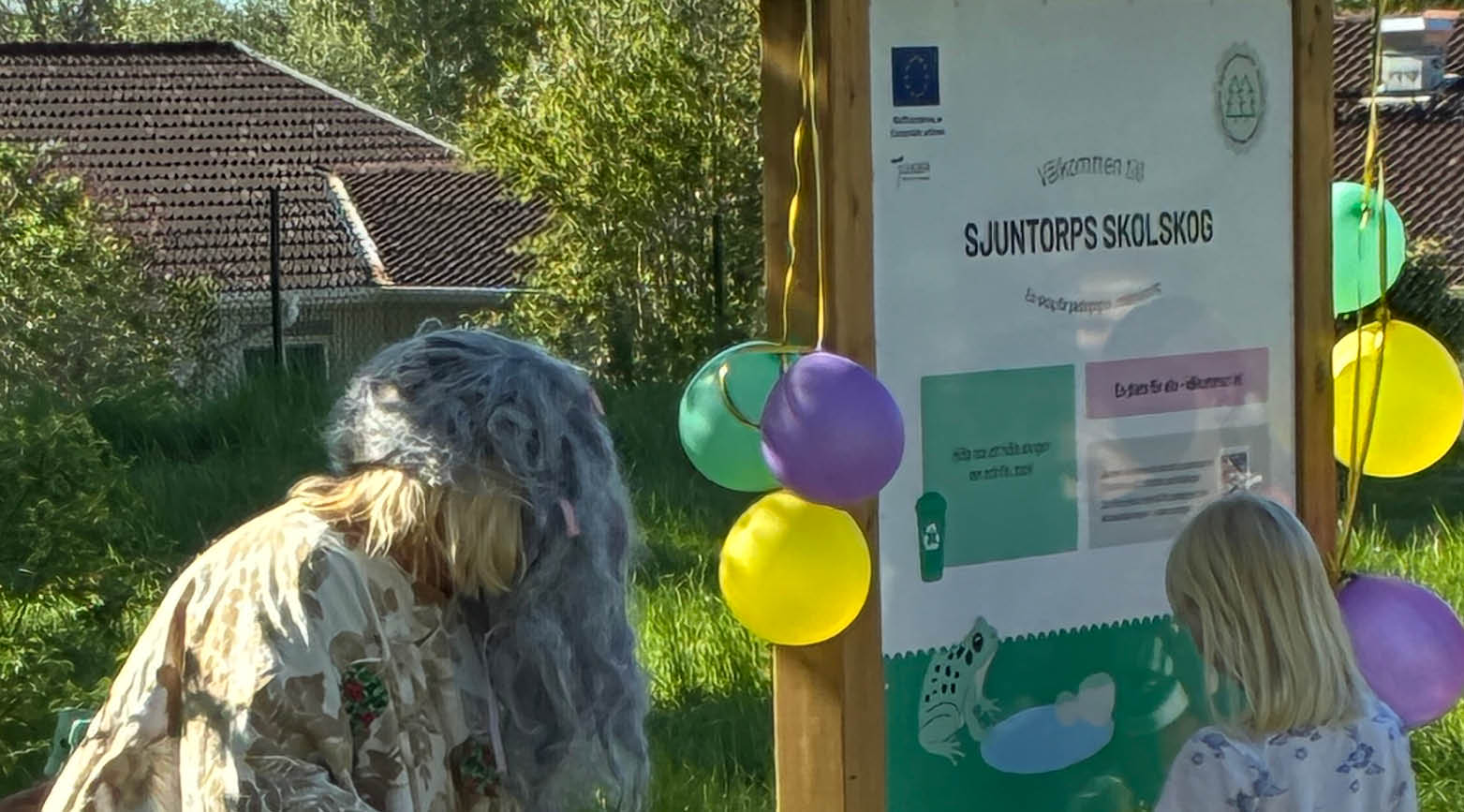 En välkomnsttavla till Sjuntorps skolskog, en kvinna utklädd till troll och ett barn som klipper band. 