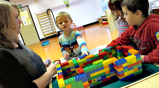 tre barn bygger ett legohus. Fröken sitter brevid och hjälper till
