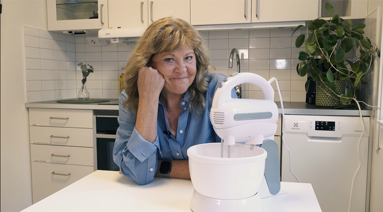 Kvinna sitter i ett kök och ser nöjd ut. På bordet står en elvisp på ett stativ och en bunke.