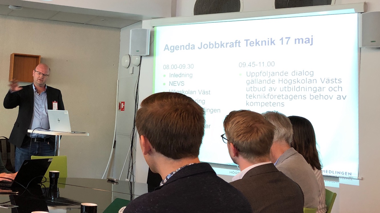 Bilden föreställer en man i grå kavaj som håller en presentation. I förgrunden sitter fyra personer och lyssnar på mannen.