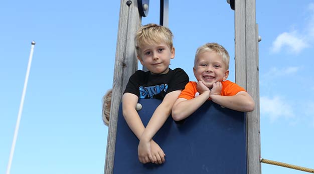 Bilden föreställer två pojkar som står uppe på en lekställning och ler stort mot kameran. Pojken till vänster i bild har en svart tröja och blont hår. Pojken till höger har en orange tröja. I bakgrunden syns klarblå himmel. 