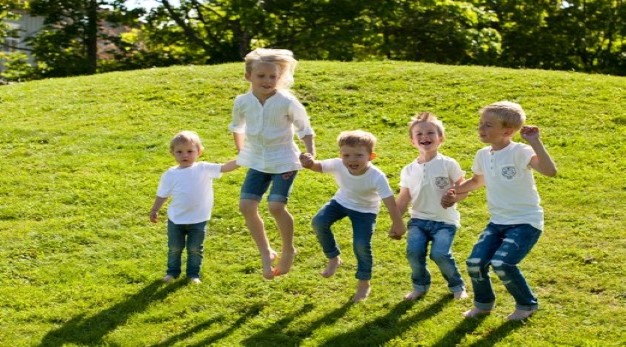 Bilden föreställer fem barn som står på rad och håller varandra i händerna samtidigt som de hoppar. Alla barnen har vita tröjor och blåa jeans. De ser alla glada ut.