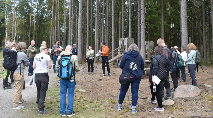 Personer i skog står i cirkel och lyssnar på en som pratar.