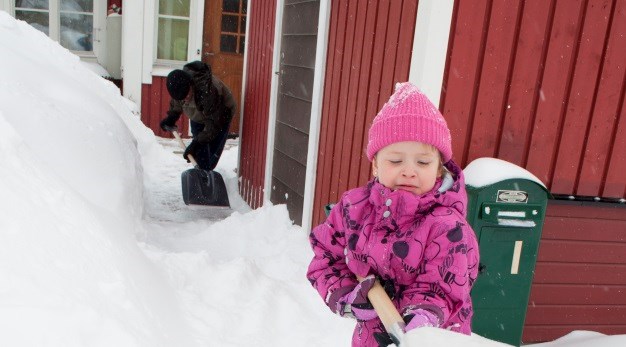 Hus omgiven med mycket snö, en vuxen och ett barn som skottar fram en gång från husets ytterdörr