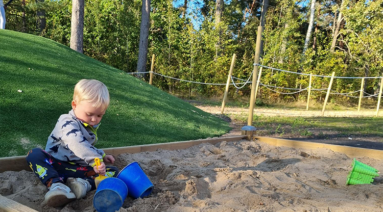 Foto från en lekplats. Ett barn sitter i en sandlåda och leker med en gul spade och två blåa hinkar. Bakom ser man skymten av en linbana.