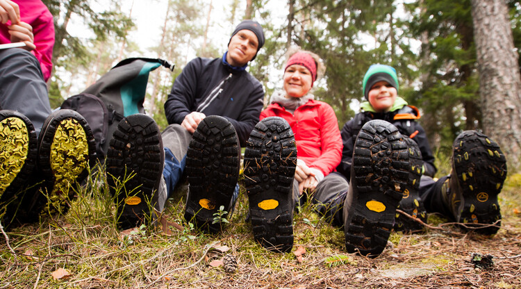 Familj sittandes i skogen, utrustade med friluftsutrustning. Skosulor i förgrunden. 