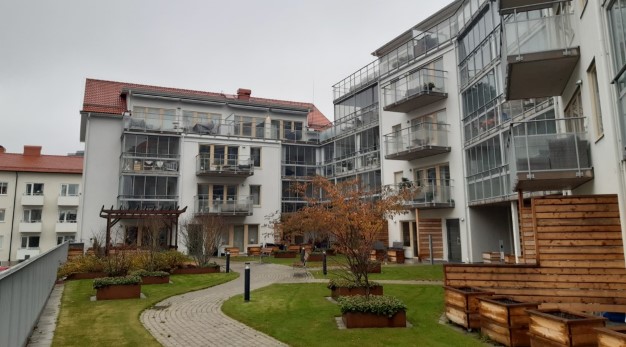 Vit fasad med stora inglasade balkonger som vetter mot flerbostadshusets innergård. Längs med innergården är det en stenlagd gång med växter och träd längs sidorna.