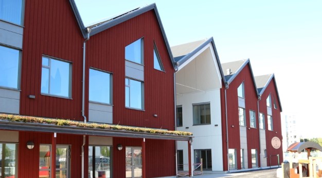 Foto av förskolans röda och vita fasad med stora glasfönster som sträcker sig hela vägen ner till golvet. Himlen är blå och i kanten av bilden skymtar man en lekplats framför förskolan.