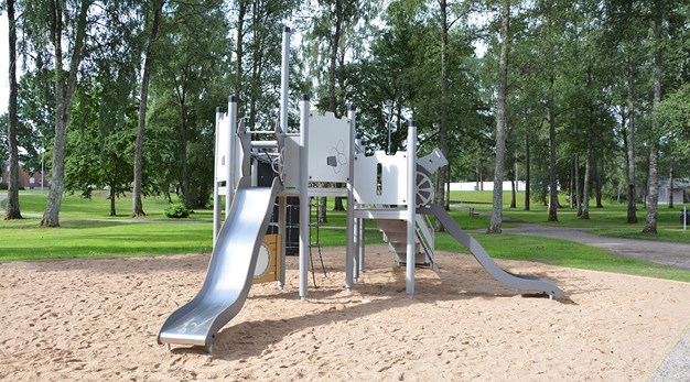 Bilden föreställer en ljusgrå och silvrig lekställning med två rutschkana. Lekställningen är formad som ett fort eller en borg. Det är sand under lekställningen och sanden lyser. Solen reflekterar i rutschkanan. 