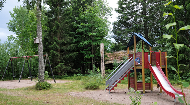 Fotot är en bild över en lekplats. I bakgrunden till vänster i bild står en grön gungställning med tre platser. Precis framför gungställningen står ett stort träd med en fågelholk. I förgrunden till höger syns en lekställning med en rutschkana. 