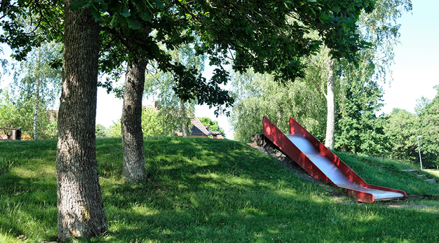 Bilden föreställer lekplatsen Björkedal. Till höger i bild är det en rutschkanan som är belägen på en gräskulle. Till vänster i bild står två stora trädstammar. 