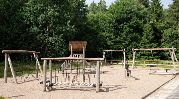 Bilden är en översiktsbild över Björndals gärdes lekplats. Från vänster i bild syns en klättervägg, en ställning för balansgång, en lekställning med en rutschkana, en gungställning med två gungor och en ställning med en kompisgunga. Bakom lekplatsen är det tät skog. På marken är det sand. Fotografiet är taget på sommaren. Solen lyser.