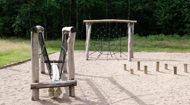 Bilden föreställer en lekplats. Till vänster i bild syns en trädfärgad ställning med balansgång. Centrerat i bild är en klättervägg som liknar ett spindelnät. Till höger i bild är flera stubbar i marken för balansgång. På marken är det sand. 