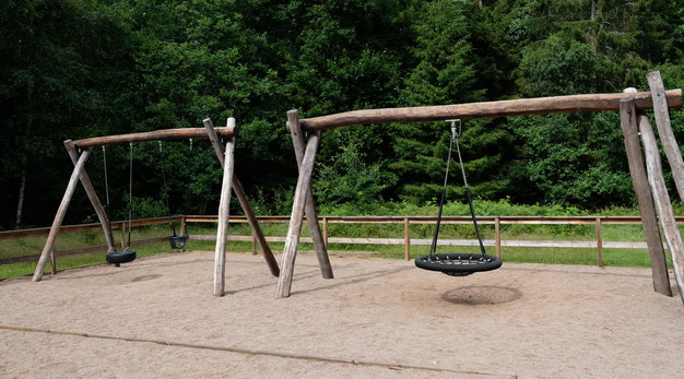 Bilden är ett fotografi över två trädfärgade gungställningar. Till vänster står en gungställning med en "däck-gunga" och en gunga för småbarn. Till höger finns en kompisgunga. Gungorna är svarta och underlagt är sand. I bakgrunden syns ett staket och en tät skog.