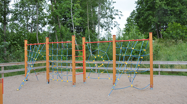 Bilden föreställer en jättelång och stor klättervägg med olika former av klättermöjligheter. Ställningen är gjord i trä och repen som man klättrar på är blåa med gula detaljer. I bakgrunden syns ett staket i trä och flera träd och buskar. 