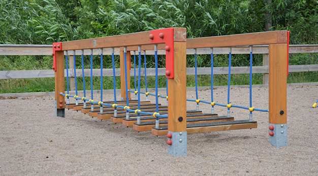 Bilden föreställer en balansbro. Bron är gjord i trä med blåa rep som löper från ställningen till balansbrädorna. Marken under är sand och i bakgrunden syns ett staket och flera gröna buskar. 