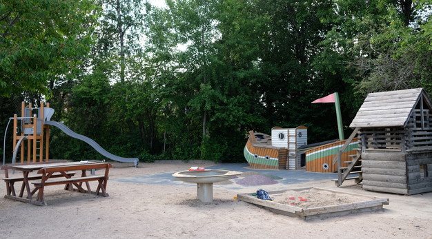 Fotografiet är en översiktsbild över lekplatsen Brunörten. Från vänster i bild syns ett parkbord i trä, en stor lekställning med en rutschkana, ett bakbord, en lekbåt, ett lekhus och en sandlåda. I bakgrunden syns mycket grönska. 