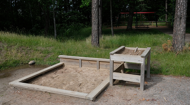 Bilden föreställer en kvadratisk sandlåda. På sandlådans högra sida står en sandlåda i bordshöjd. I bakgrunden syns två träd och en vildvuxen gräsplätt. 