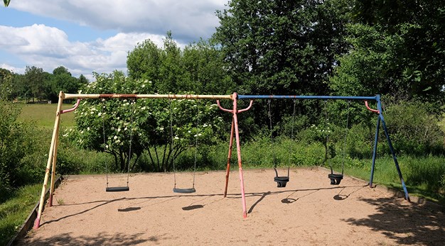 Bilden föreställer en gungställning i gult, blått och rött. Gungställningen har fyra gungor varav två är för små barn. Marken under är sand och runt omkring lekplatsen är det mycket grönska. 