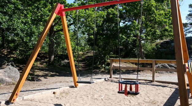 Bilden föreställer en gungställning med två gungor. Gungställningen är i trä och har röda detaljer. I bakgrunden syns gröna träd. Marken är sand. Fotografiet är taget en solig dag. 