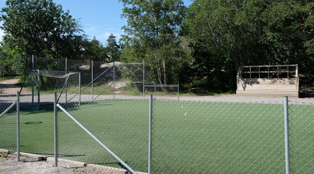Bilden är ett foto av en del av en fotbollsplan. I förgrunden runt fotbollsplanen är det ett grått staket. I bakgrunden finns det en läktare i trä