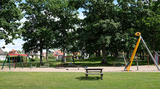 Bilden är en översiktsbild över hela Ekparken. Från vänster i bild syns följande: en gungställning, en kompisgungar, en cykelkarusell, gungbräda, parkbord, klätterpyramid och en linbana. I bakgrunden syns stora gröna träd. 