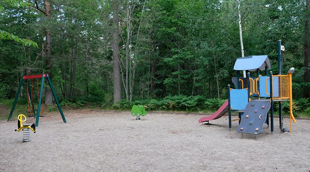 Fotografiet är en översiktsbild över en lekplats. Från vänster i bild sysn ett gult dgungdjur, en röd och grön gungställning, ett grönt gungdjur och till sits en lekställning med rutschkana och klättervägg. Marken är sand och i bakgrunden syns mycket grönska. 