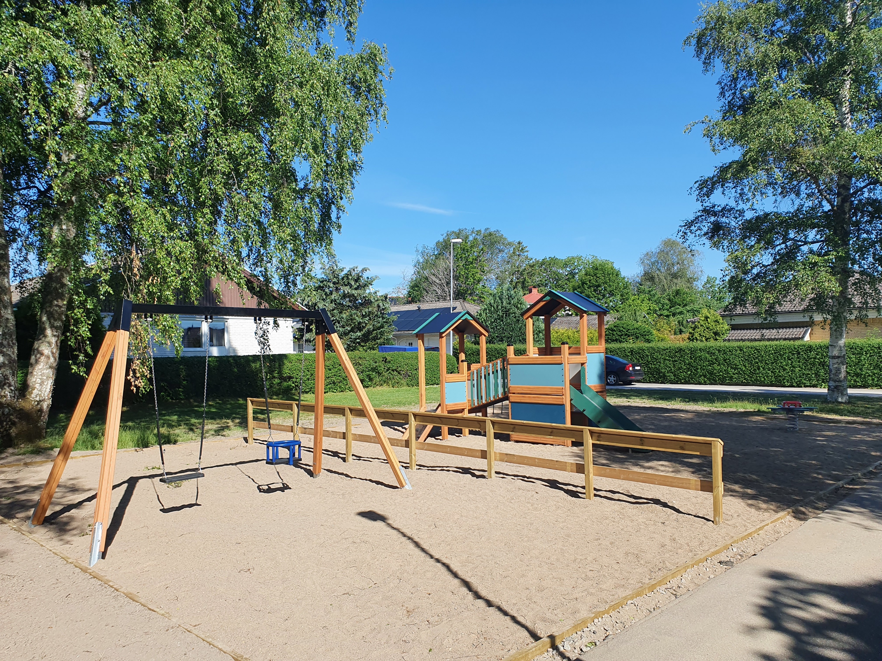 Fotografiet är en översiktsbild över Fågelparkens lekplats. I förgrunden syns en röd cykelkarusell. I bakgrunden finns det en röd gungställning. Marken i lekplatsen är sand och runt omkring är det gräs.