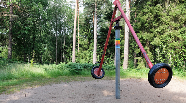 Bilden föreställer en mobilusgunga. Gungan är röd och marken under är sand. I bakgrunden syns ett skogsparti