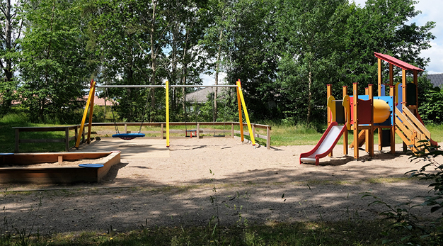 Bilden är en översiktsbild över Karolinerparkens lekplats. Från vänster i bild är det en sandlåda, en gul gungställning och en stor lekställning med två rutschkanor. Marken under är sand. 