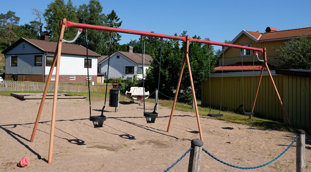 Bilden föreställer en gungställning med fyra platser. Gungorna längst till vänster är gungor för små barn och de till höger är för stora barn. Ställningen är röd och orange. I bakgrunden syns två vita hus med brunt tak. Marken är sand. På marken syns skuggor från lekställningen. 