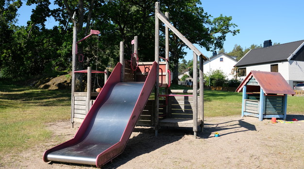 Bilden föreställer en liten lekställning med en rutschkana. Rutschkanan är röd och bred. I bakgrunden till höger syns ett litet blått lekhus med rött tak. Marken är sand och i bildens högra hörn syns ett vitt hus med svart tak. 