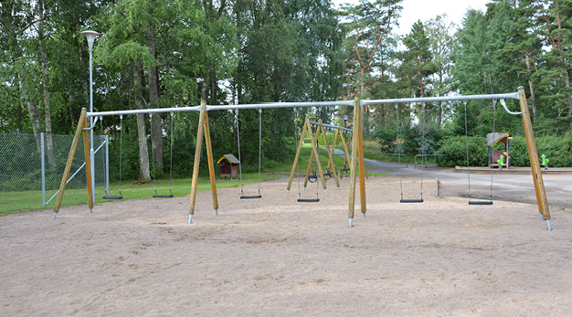 Bilden är ett foto av en gungställning med sex stycken gungor. Till vänster om ställningen, som är i trä och silver, står en lyktstolpe i silver. Bakom den är det flertalet träd och det ger lekplatsen en mysig känsla. 