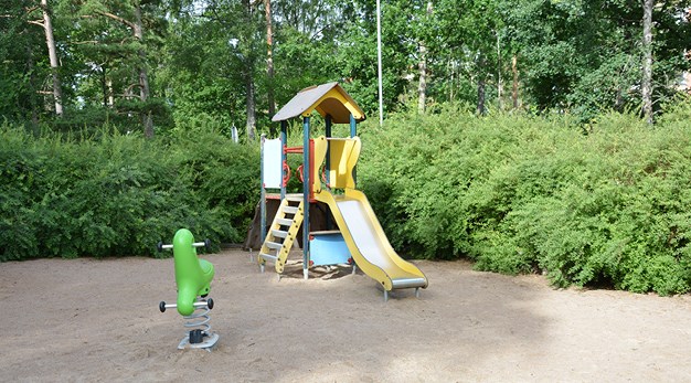 Bilden föreställer en lekställning som står alldeles intill en jättestor buske. Lekställningen är gul, röd och blå och har en rutschkana. Framför lekställningen står ett grönt gungdjur.  