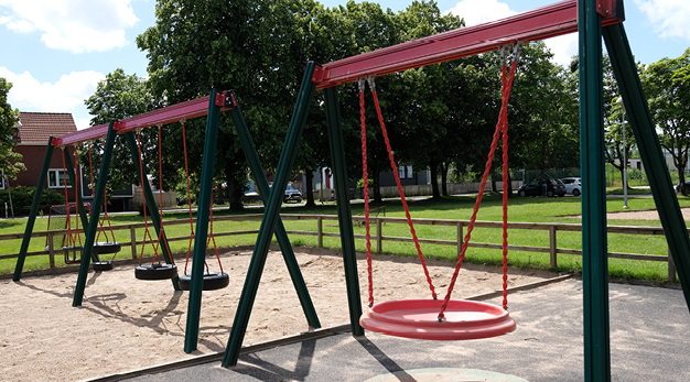 Bilden föreställer två gungställningar i grönt med röda detaljer. Gungorna är omringade av ett staket. På marken är det sand. På gungställningen längst bort i bild är det fyra däck-gungor. På ställningen närmast i bild är det en röd kompisgunga. 