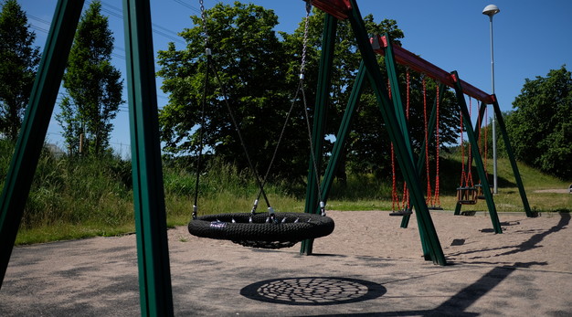 Bilden föreställer en svart kompisgunga som sitter på en grön ställning. I bakgrunden syns flera gungor och gungställningar. I bakgrunden syns blå himmel och trädkronor. 